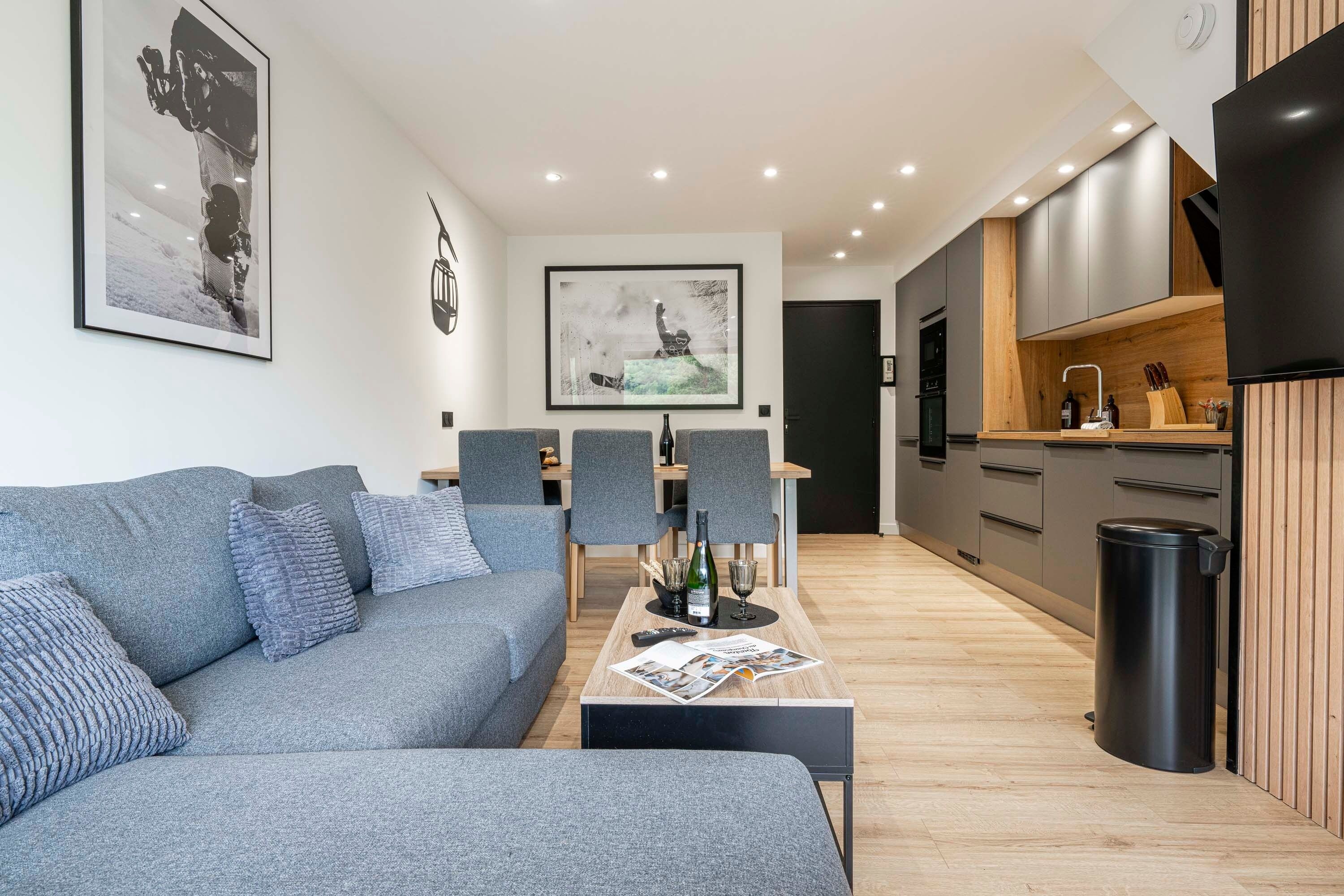 A modern living room with a grey sofa, a coffee table with a magazine, and a kitchen area with grey cabinets and a black trash bin.