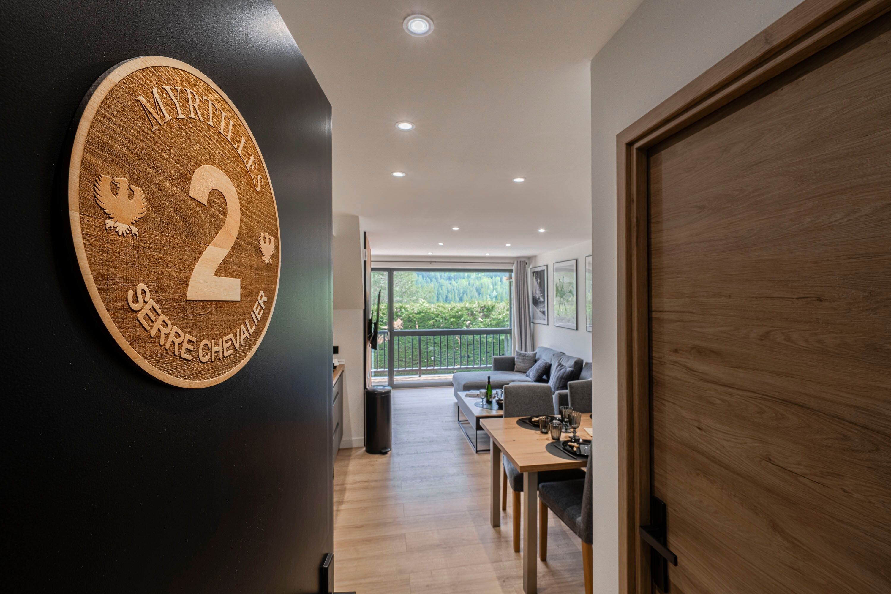 A modern interior with a wooden door, a round plaque with 'MYRTILLES 2' and a crest, a balcony with a view, and a seating area with a table.