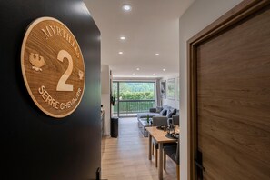 A modern interior with a wooden door, a round plaque with 'MYRTILLES 2' and a crest, a balcony with a view, and a seating area with a table. - Suite Myrtilles - Bel appartement moderne pour 4 (Saint-Chaffrey)