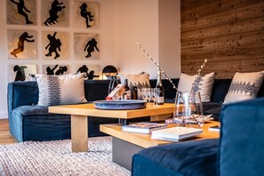 A modern living room with a wooden coffee table, a sofa, and a rug. There are books, a bottle of wine, and a vase with branches on the table. - Malago - Five Bedroom Chalet, Sleeps 14 (Le Monêtier-les-Bains)