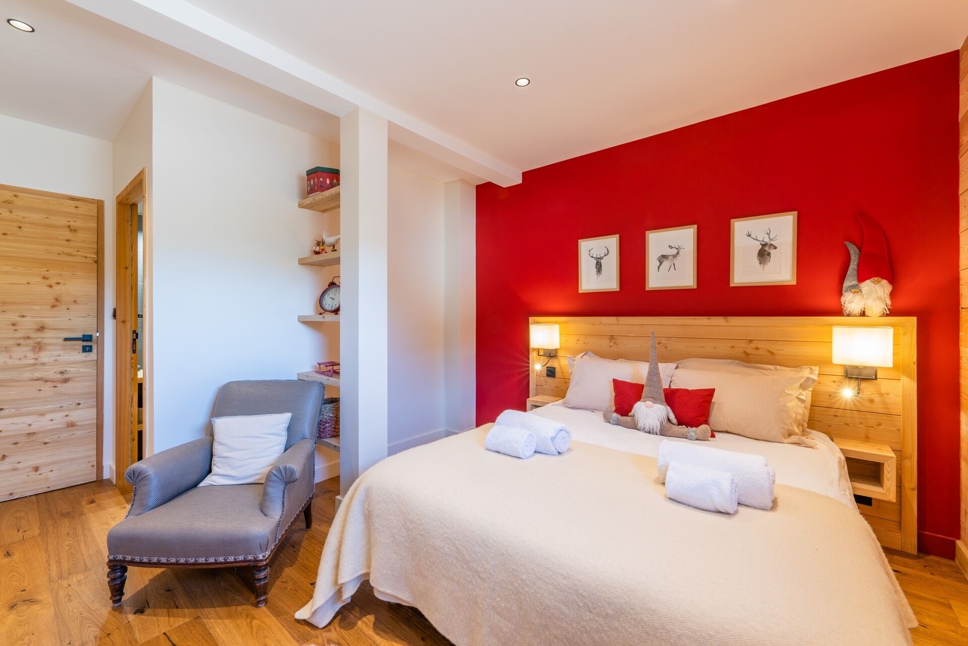Une chambre avec un mur rouge, une tête de lit en bois, un lit avec du linge de maison blanc, un fauteuil gris et une étagère avec des objets décoratifs.