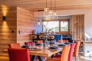 A dining area with a wooden table set for four, surrounded by red chairs, with hanging pendant lights above. - Malago - Five Bedroom Chalet, Sleeps 14 (Le Monêtier-les-Bains)
