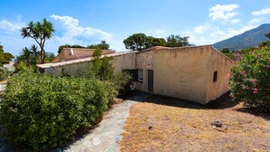 A rustic building with a stone pathway leading to a door, surrounded by greenery and a mountain in the background. - Le Cocody - Appt avec terrasse, proche de la plage (Lumio)