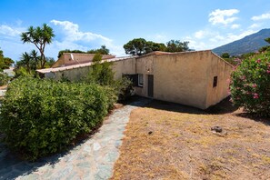 A rustic building with a stone pathway leading to a door, surrounded by greenery and a mountain in the background. - Le Cocody - Appt avec terrasse, proche de la plage (Lumio)