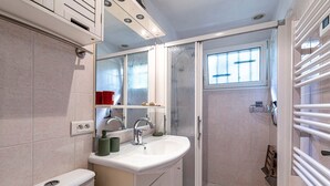 A bathroom with a white sink, mirror, and toilet, a shower with a glass door, and a window providing natural light. - Le Cocody - Appt avec terrasse, proche de la plage (Lumio)
