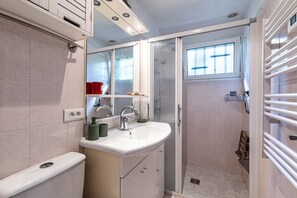 A bathroom with a white sink, mirror, and toilet, a shower with a glass door, and a window providing natural light. - Le Cocody - Appt avec terrasse, proche de la plage (Lumio)