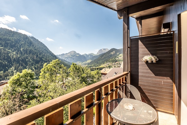 A balcony with a view of mountains, a wooden railing, and a table with a chair.