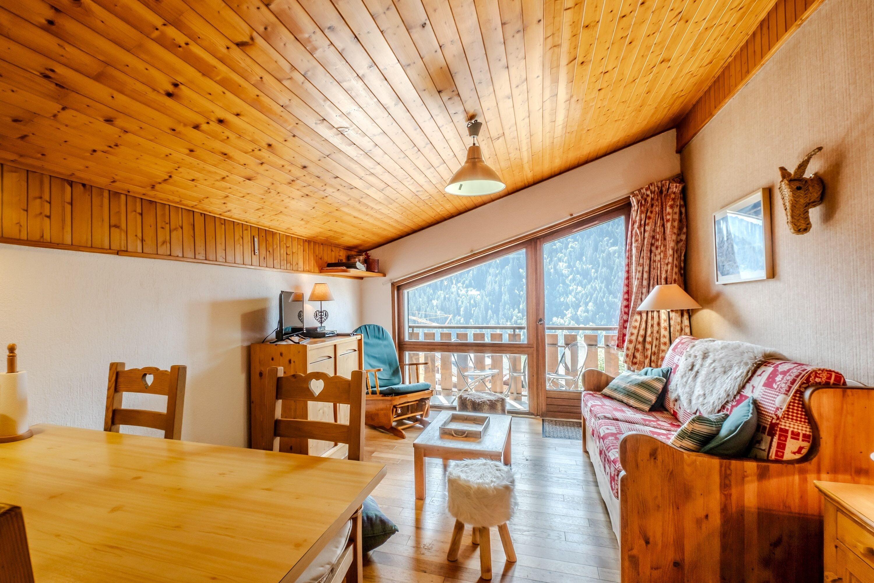 A wooden interior with a dining table, chairs, a sofa, and a view through a large window.