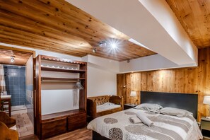 Ein Schlafzimmer mit Holzboden, einem Bett mit gemustertem Überwurf, einem Nachttisch, einer hölzernen Bank und einem Badezimmer mit blauen Fliesen.
