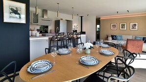 A modern dining area with a wooden table set for four, black chairs, and a kitchen in the background. - Ker Siméon - Maison moderne à 600 m de la plage (Saint-Cast-le-Guildo)