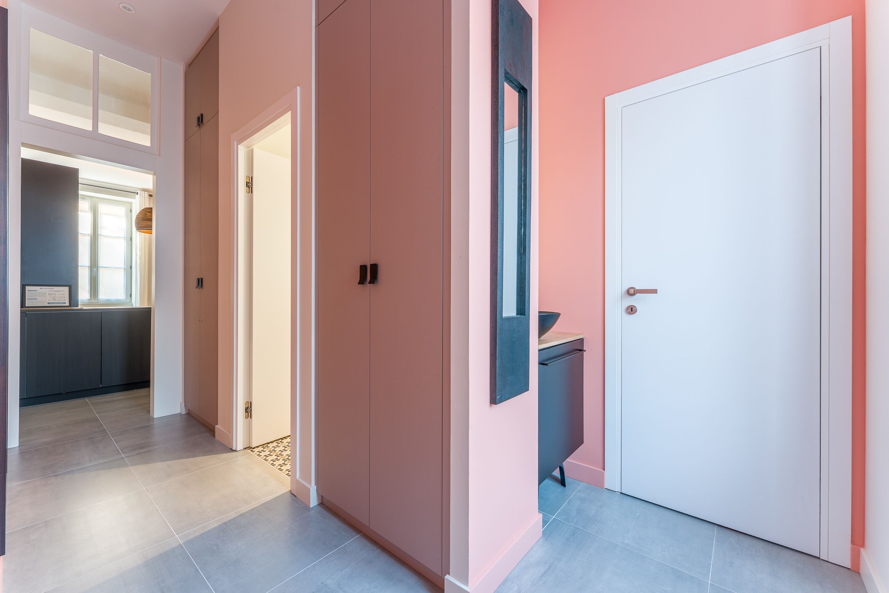 A hallway with a door, a mirror, and a kitchen area in the background.