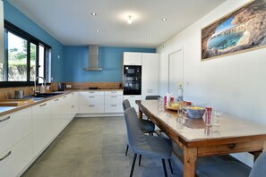 Une cuisine moderne avec des meubles blancs, une table à manger en bois et une vue sur la mer par la fenêtre.