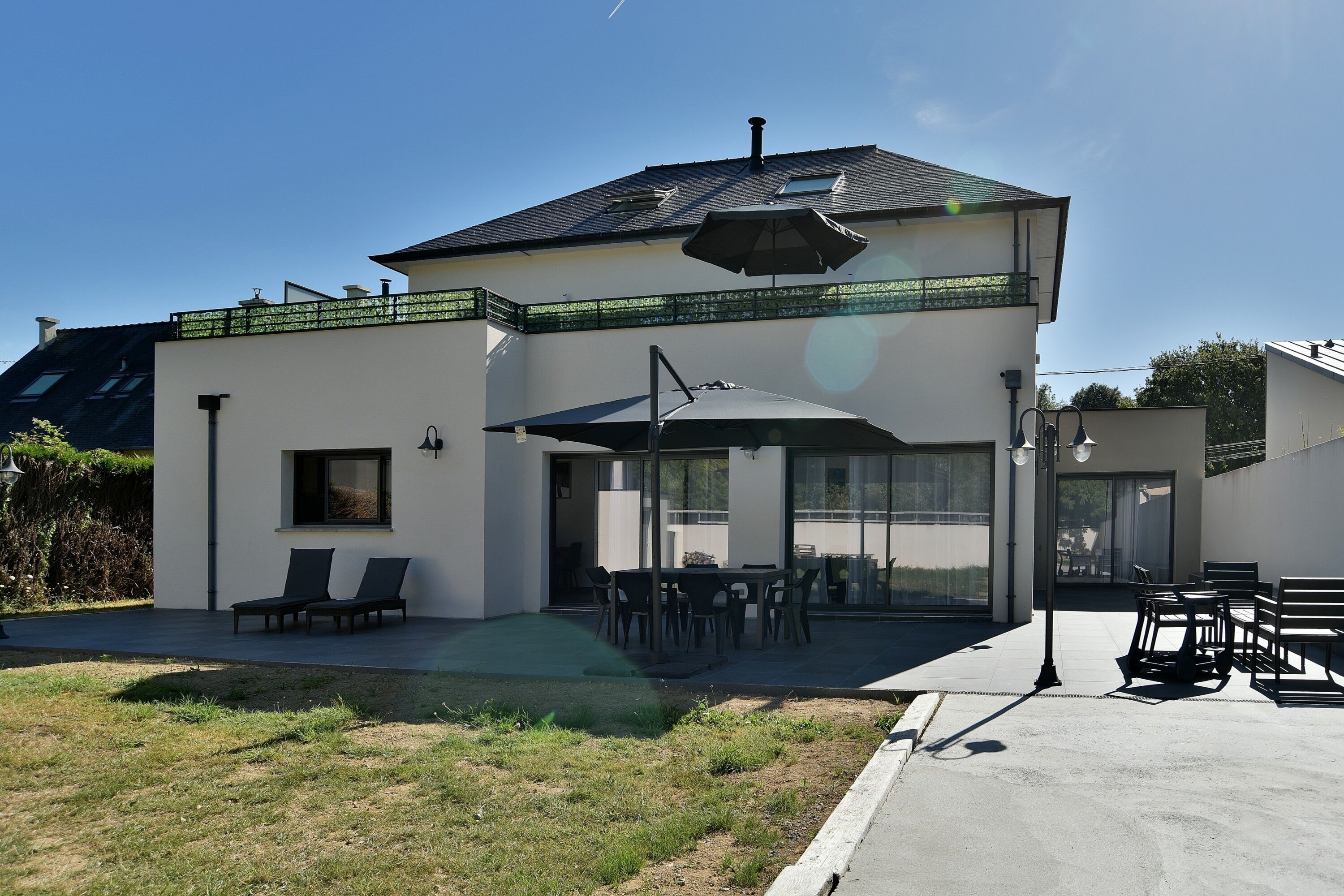 Ein modernes, zweigeschossiges Haus mit Terrasse, Sitzgelegenheiten im Freien und einem überdachten Bereich.