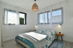 Ein Schlafzimmer mit einem Bett, zwei Nachttischen, einer Hängelampe und zwei Fenstern mit transparenten Vorhängen.