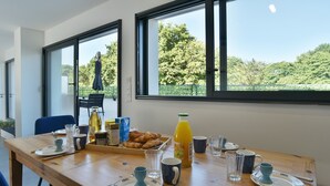 A breakfast table set with plates, cups, and a bottle of orange juice, with a view of trees through large windows. - Ti Kastrilhez - 1er - À 1 km de la plage (Saint-Cast-le-Guildo)