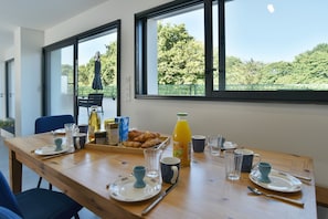 A breakfast table set with plates, cups, and a bottle of orange juice, with a view of trees through large windows. - Ti Kastrilhez - 1er - À 1 km de la plage (Saint-Cast-le-Guildo)