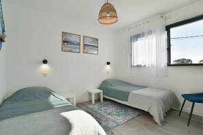 A bedroom with two beds, a small table, a chair, a window with curtains, and two wall-mounted lights. - Ti Kastrilhez - 1er - À 1 km de la plage (Saint-Cast-le-Guildo)