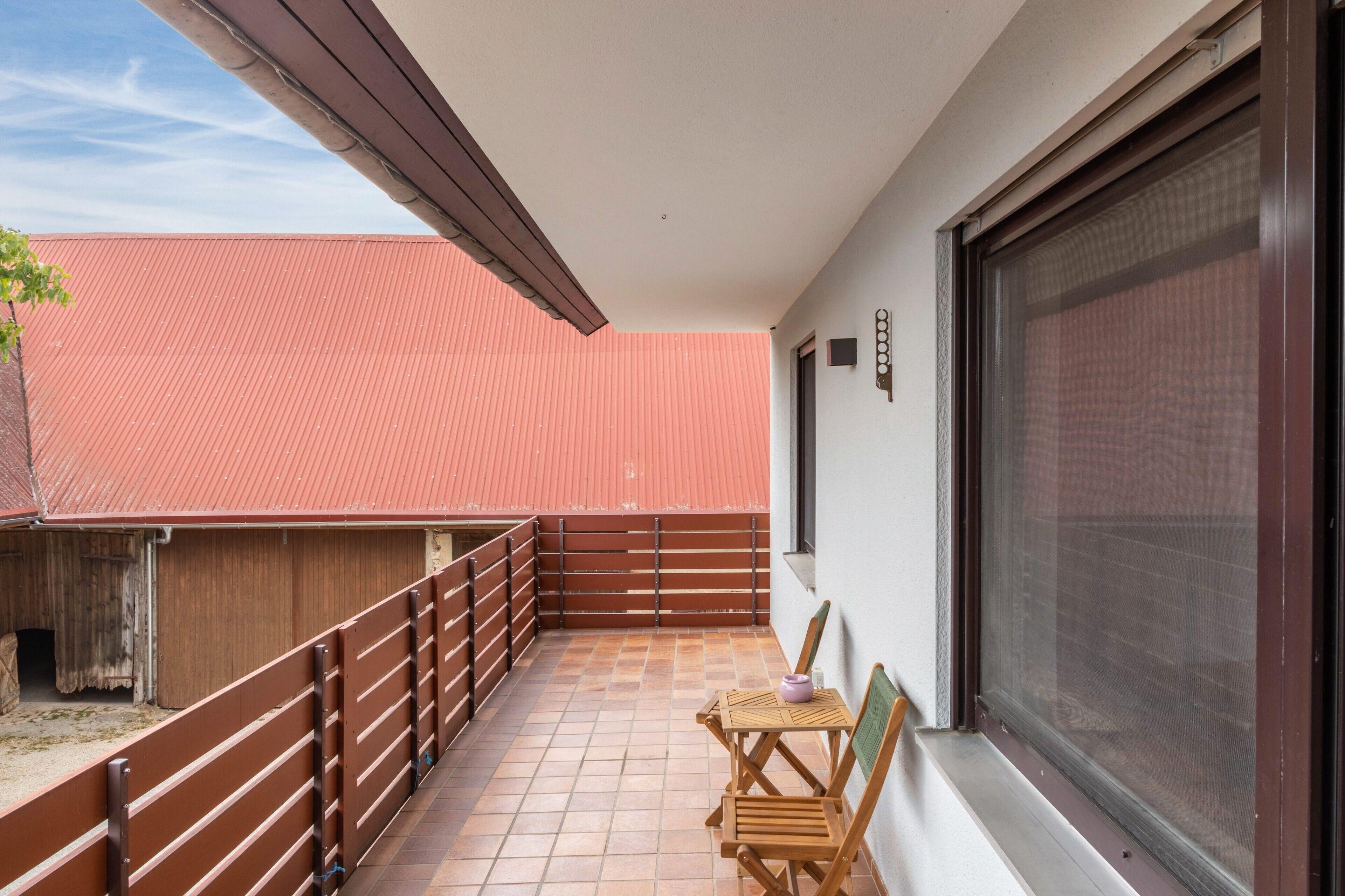 Ein Balkon mit Blick auf ein rotdaches Gebäude, ein Holzstuhl und ein Tisch mit einer Vase.