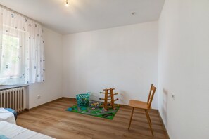 Una habitación con piso de madera, una sola silla de madera, una mesita, un radiador y una ventana con cortina.