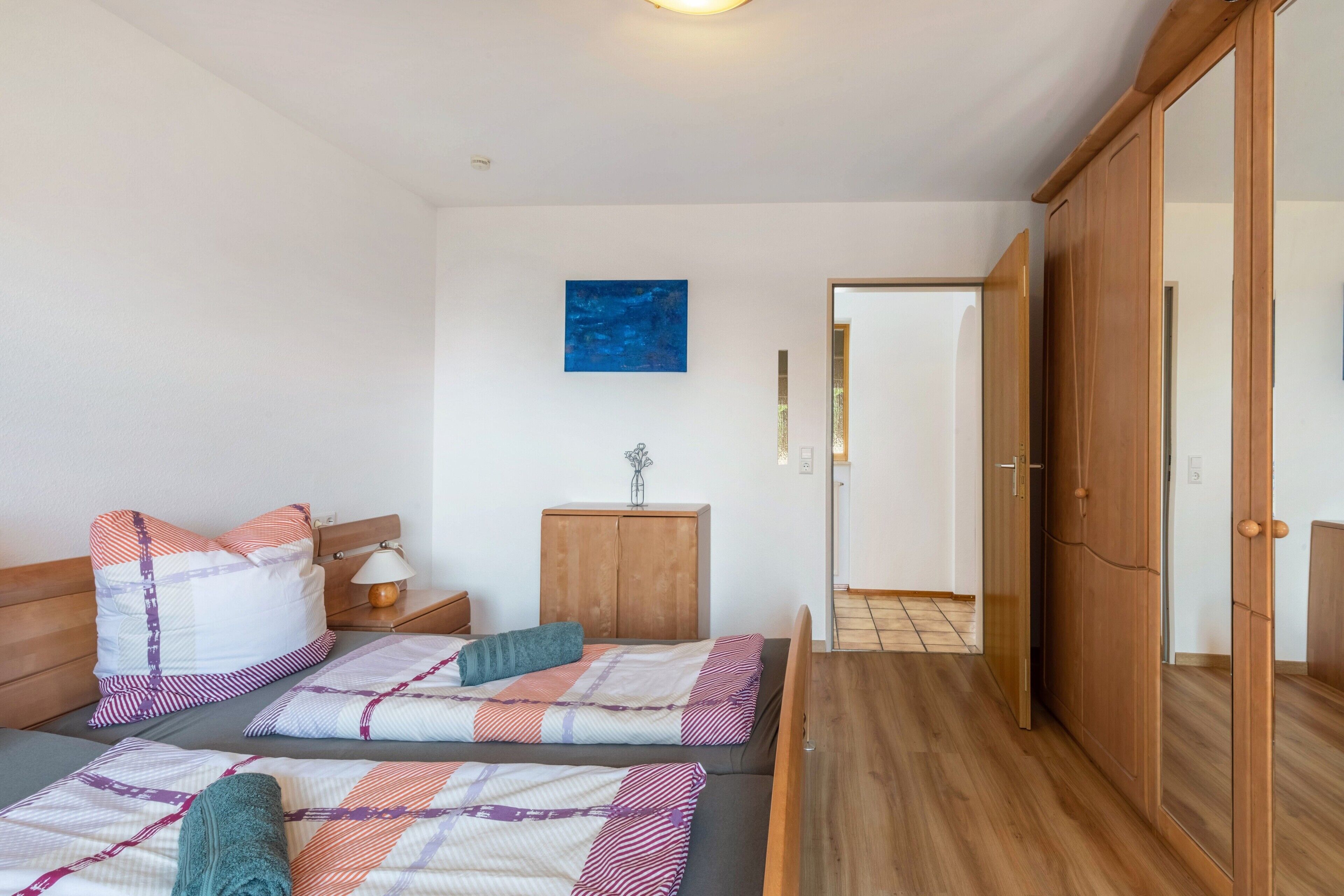 Ein Zimmer mit zwei Betten, einem Holzschrank, einem Nachttisch, einer Lampe und einem blauen Bild an der Wand.