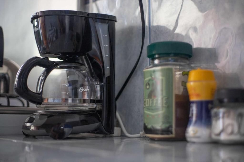 Una cafetera sobre una encimera, con un tarro de café y botellas de condimentos cerca.