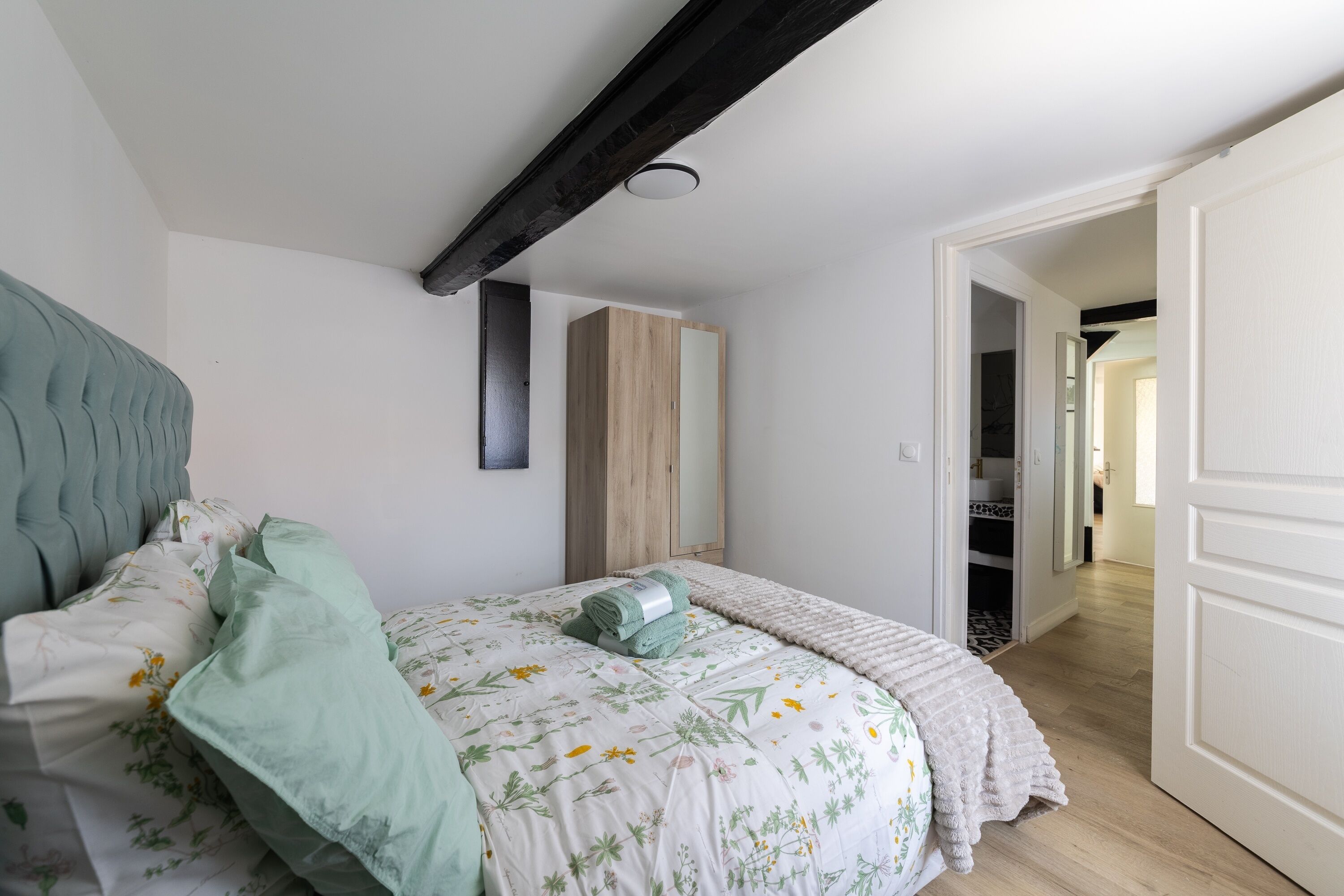 Une chambre à coucher comprenant un lit, une tête de lit, une armoire en bois, un miroir et une porte donnant sur une autre pièce.