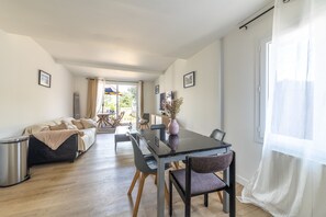 A modern living room with a sofa, dining table, chairs, and a view through a glass door to an outdoor area. - Chez Léa - Maison familiale pour 6 avec jardin (Berville-sur-Mer)