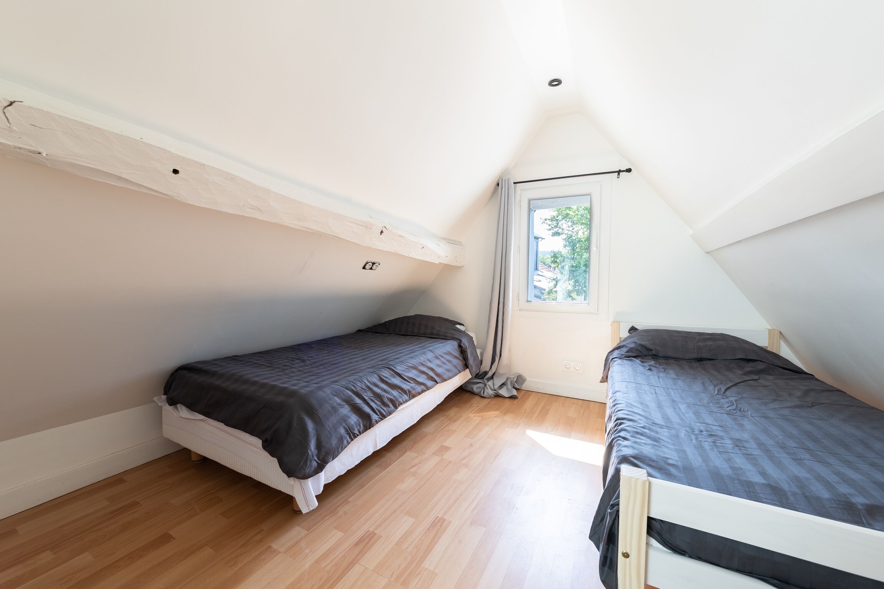 A small, single-room apartment with two beds, wooden flooring, a sloped ceiling, and a window with a view of greenery.