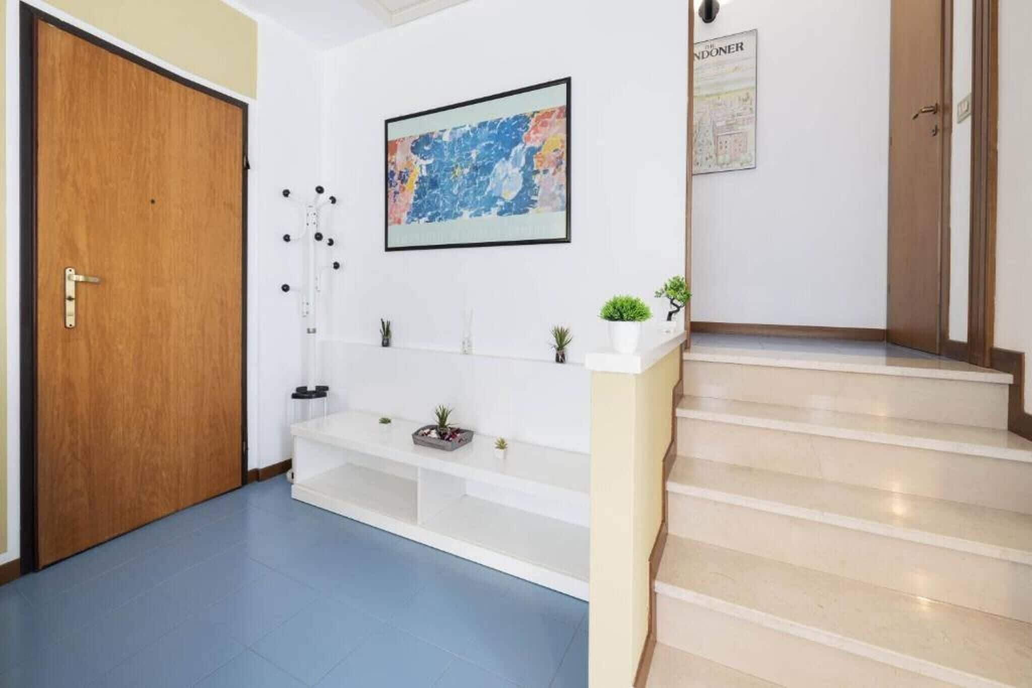 A hallway with a wooden door, a white shelf with plants, a framed abstract painting, and a staircase.
