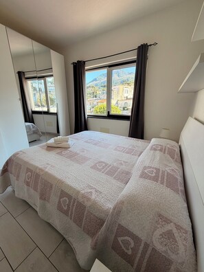 A bedroom with a bed, a window with curtains, a view of buildings, and a tiled floor.