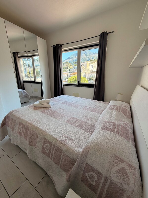A bedroom with a bed, a window with curtains, a view of buildings, and a tiled floor.