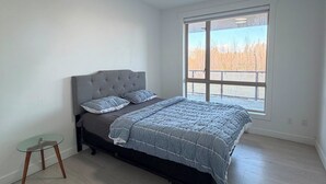 Quarto com cama, mesinha, janela com vista e luminária no teto.