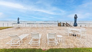 Une terrasse au bord de la plage, avec des chaises longues et des tables blanches, un escalier en bois menant à la plage et une vue sur l’océan.