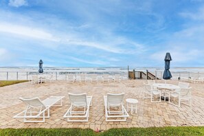 Eine Strandpatio mit weißen Liegestühlen und Tischen, eine Holztreppe zum Strand und Blick aufs Meer.