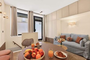 Ein Hotelzimmer mit Esstisch, gedeckt mit Früchten, einem Croissant und Orangensaft. Im Raum befinden sich eine Couch und ein Sessel.