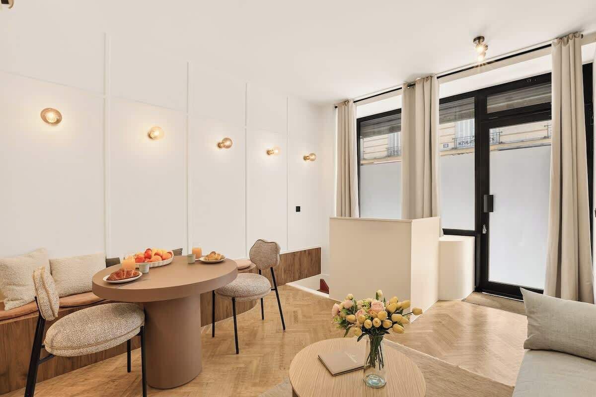 A modern hotel room with a round dining table, a sofa, and a view of the city through large windows.