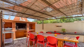 An outdoor patio with a wooden table, red chairs, a grill, and a stone floor.