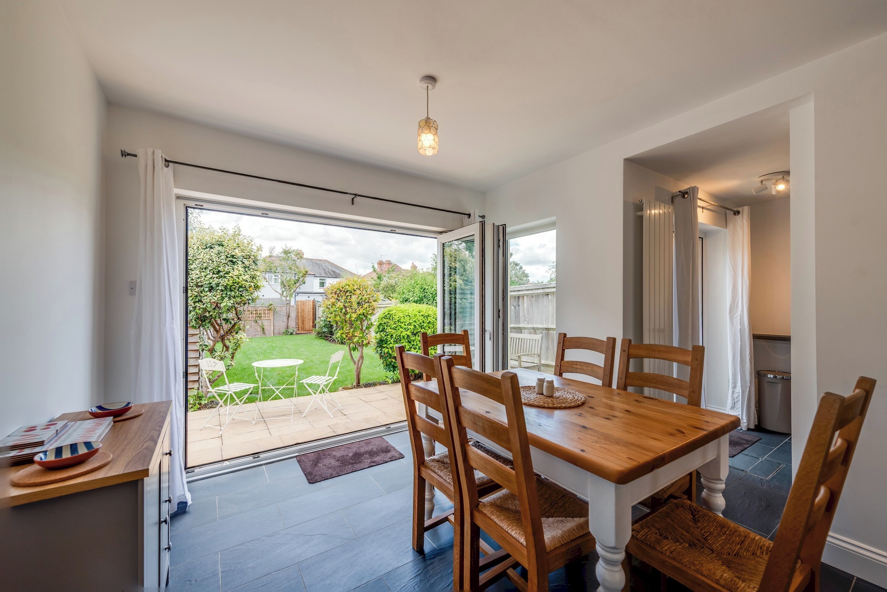 Un coin repas avec une table et des chaises en bois, une vue sur un jardin à travers des portes vitrées et un luminaire au plafond.