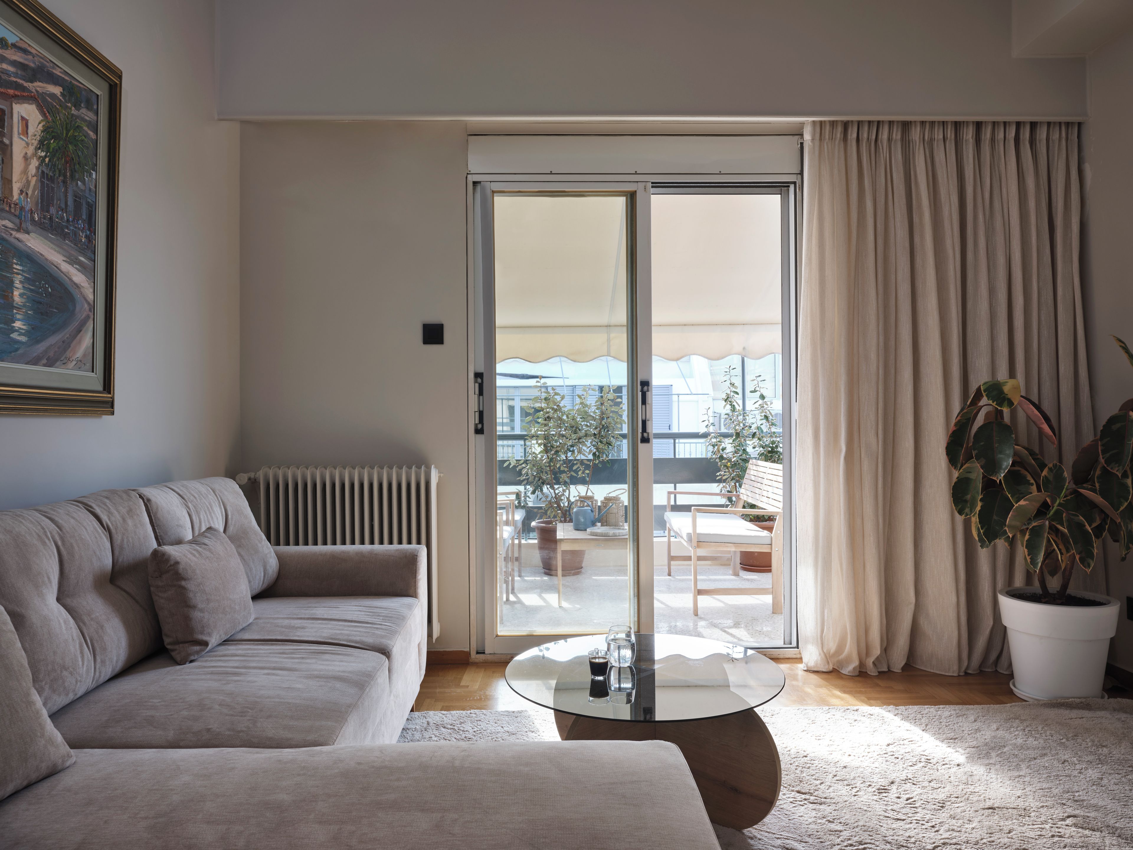 A living room with a sofa, a glass coffee table, a radiator, a sliding glass door leading to an outdoor area with a table and chairs, and a potted plant.