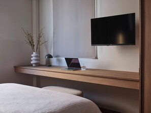 A modern hotel room with a wooden desk, a laptop, a glass of water, a vase with branches, a lamp, and a flat-screen TV mounted on the wall. - Two Bedroom Apartment (Athina)