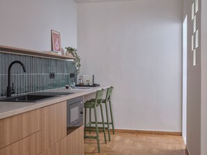 A modern kitchen with a built-in microwave, a sink, and a wooden countertop. There are two green chairs and a small table with a plant and a framed picture on the counter. - Xenios Athens Stay – 2 Bedroom Apartment (Athina)