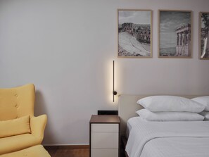 A bedroom with a bed, a nightstand, a chair, and wall-mounted artwork. - Xenios Athens Stay – 2 Bedroom Apartment (Athina)