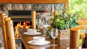 Ein Holztisch ist mit weißen Schüsseln und Weingläsern gedeckt, im Hintergrund brennt ein Kaminfeuer und ein Topf mit Grünpflanzen steht daneben.