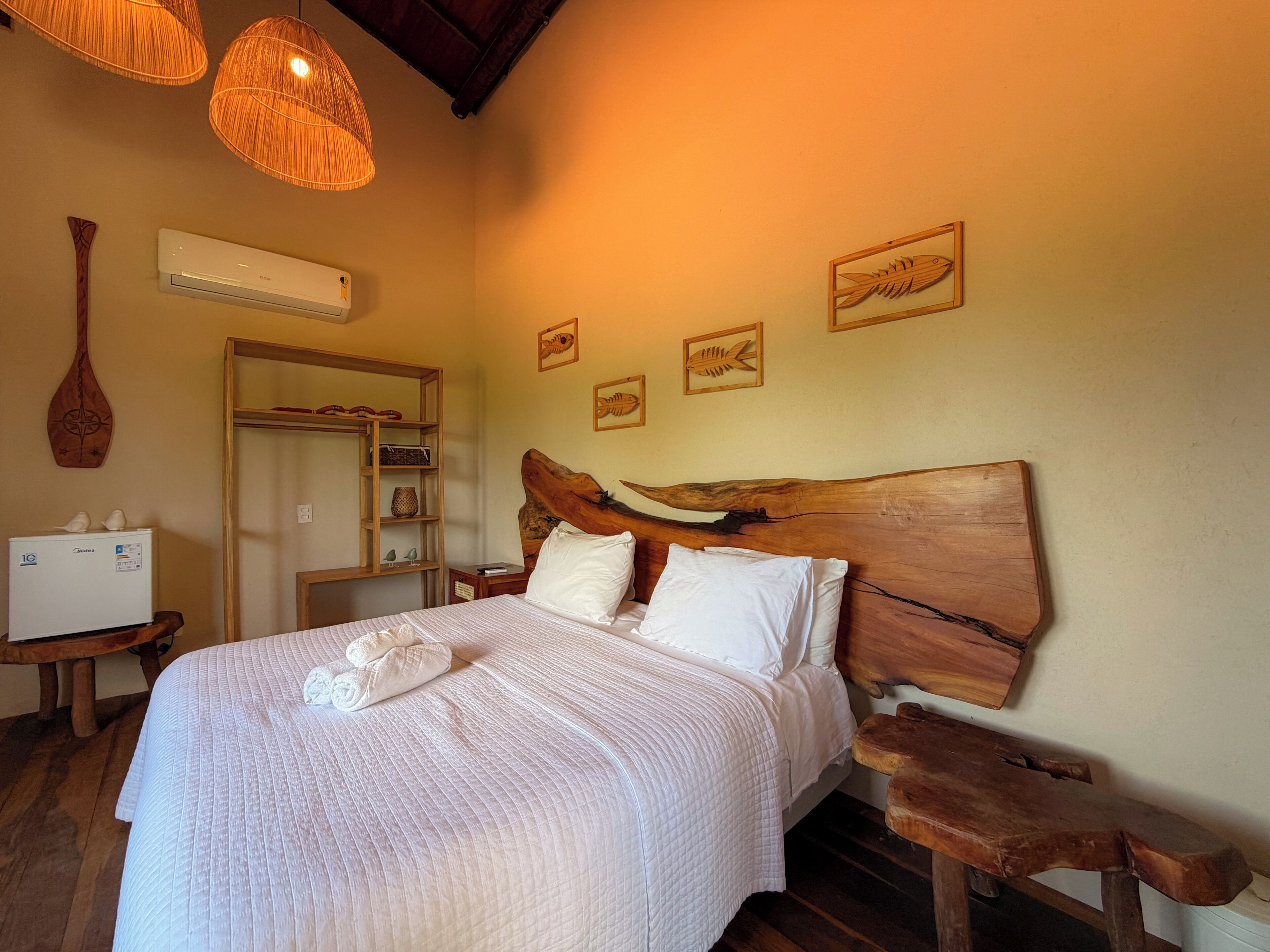 A bedroom with a wooden headboard, a bed with white bedding, a wooden nightstand, a wall-mounted air conditioner, and a wooden shelf with decorative items.