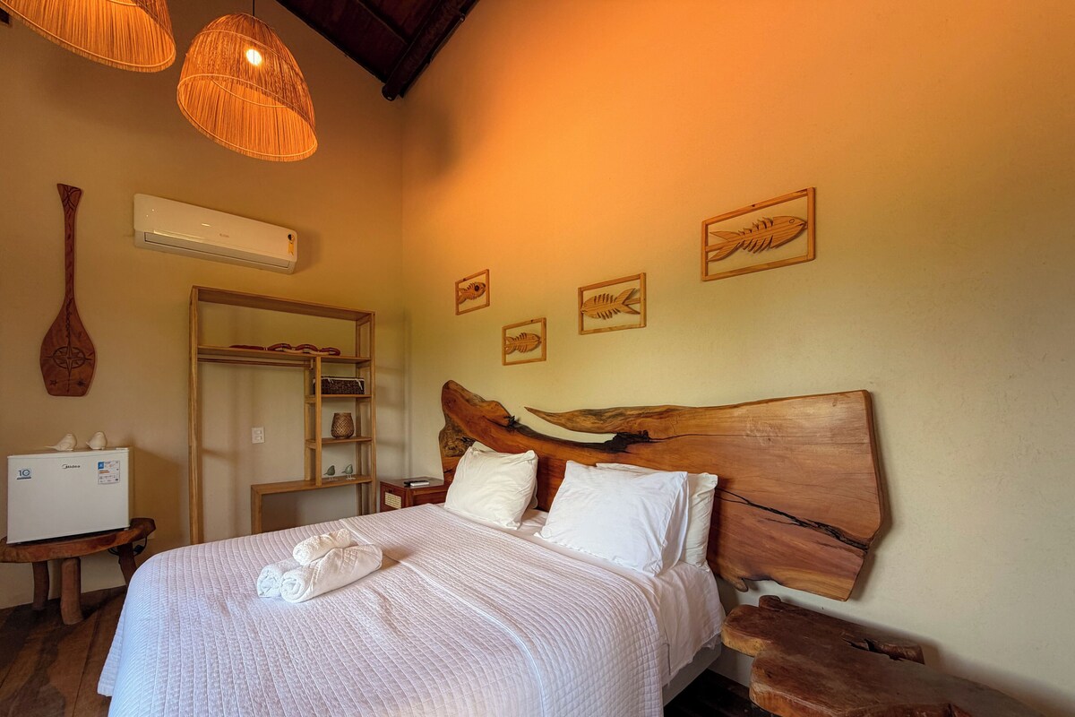 Un dormitorio con cabecera de madera, una cama con ropa blanca, una mesita de noche de madera, un aire acondicionado de pared y un estante de madera con objetos decorativos.