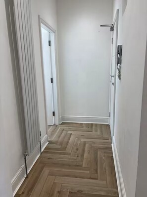 Un passadís amb sòl de fusta en relleu, parets blanques i una porta amb mànec negre.