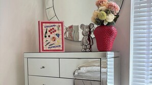 Een witte dressoir met laden, een spiegel, een teddybeer, een ingelijste foto en een vaas met bloemen.