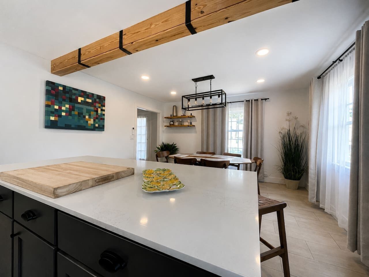 Een moderne keuken met een groot kookeiland, houten balken in het plafond, inbouwverlichting en een eethoek met een plant.