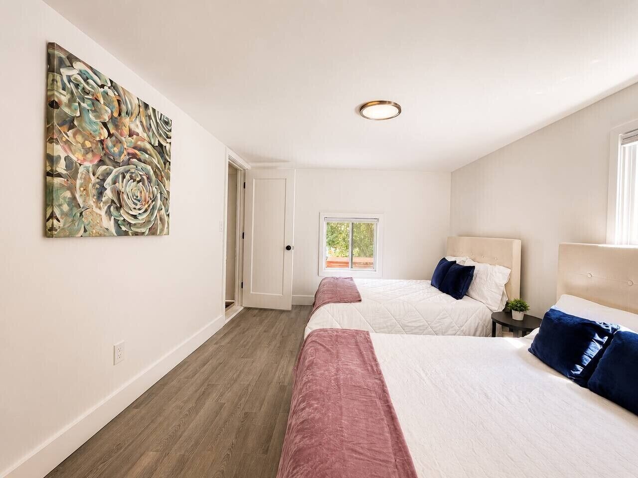 Habitación de hotel con dos camas, un cuadro abstracto grande, una ventana y una puerta que da a otra habitación.