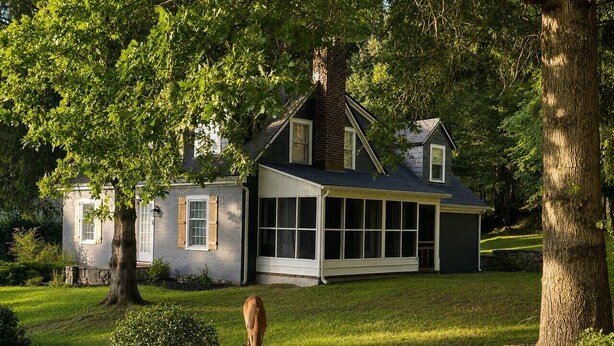 Uma casa de dois andares com uma varanda grande e envidraçada, cercada por árvores e um gramado bem cuidado.